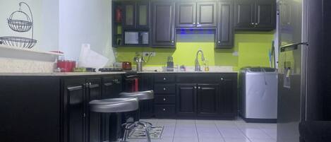 A kitchen with dark cabinets, a stainless steel refrigerator, a microwave, and a sink. There are two metal stools and a patterned rug on the floor.