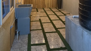 A backyard with a stone and grass floor, a wooden fence, a water tank, and string lights. - Townhouse426 gated community (Portmore)