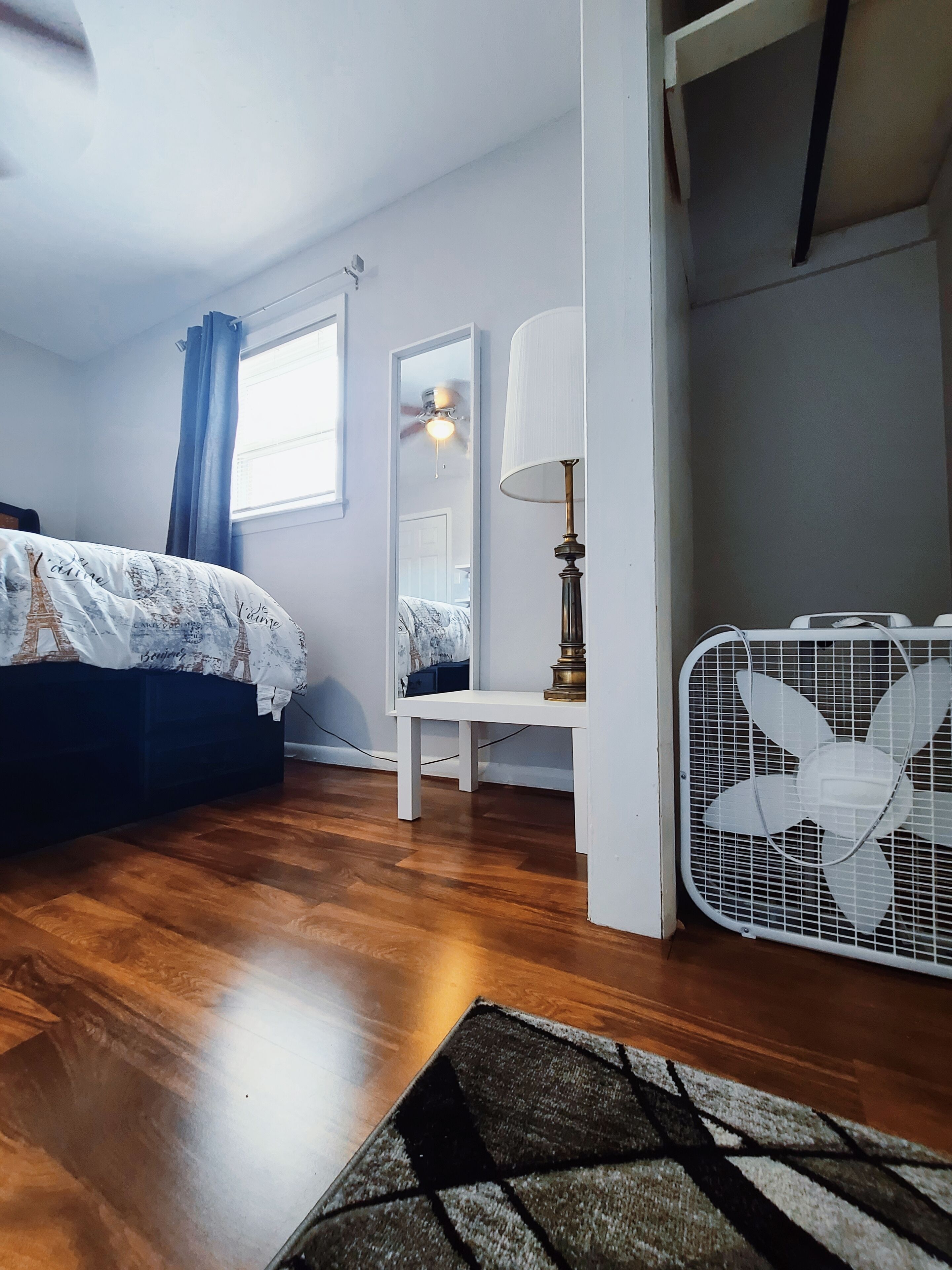 Un dormitorio con una cama, una mesita de noche con lámpara, un espejo, un ventilador de techo y un ventilador blanco en el suelo.