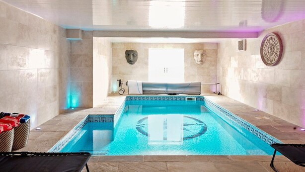 A modern indoor swimming pool with a clear blue water feature, stone walls, and a ceiling with a geometric design. - Stunning Coastal Retreat - Indoor Pool - Sauna (Hunmanby)