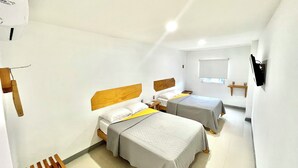 Una habitación pequeña y luminosa con dos camas, cabecera de madera, mesita de noche, televisor montado en la pared y ventana con persianas.