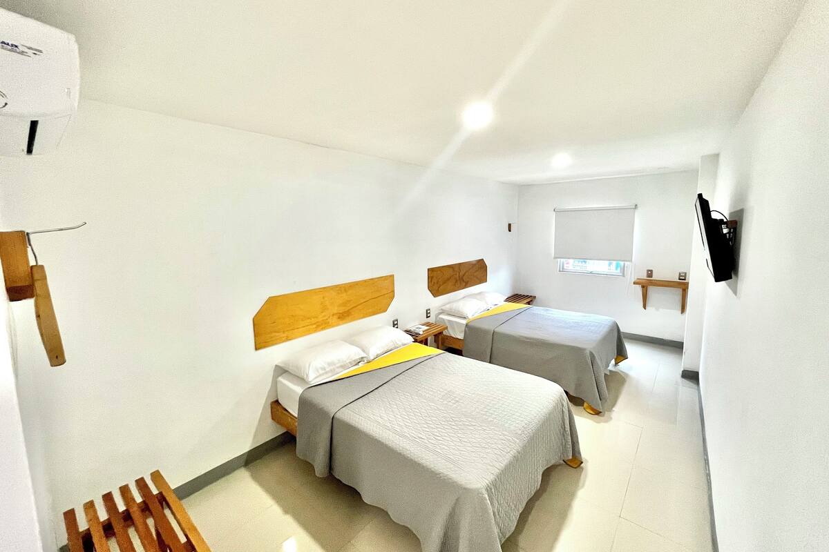 A small, well-lit room with two beds, a wooden headboard, a bedside table, a wall-mounted TV, and a window with blinds.