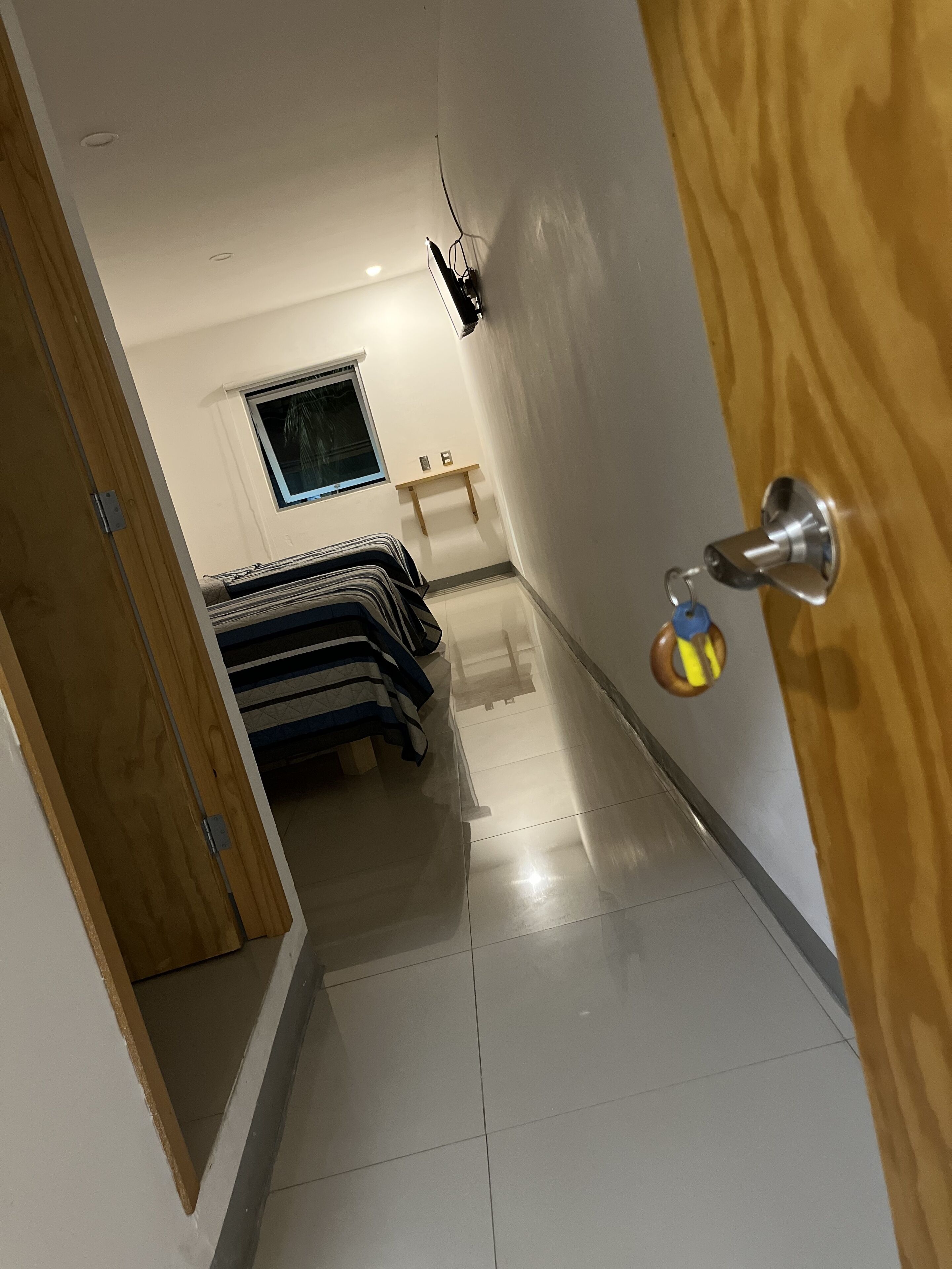 Un pasillo estrecho con una cama, un ventanal pequeño y una puerta con un llavero.