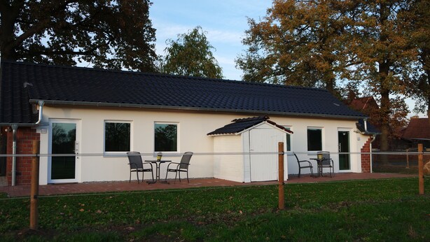 Een laag gebouw met een pannendak, een klein tuinhuisje en een omheind terras met buitenmeubilair.