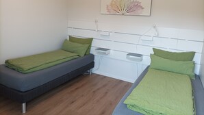 Een kleine, schone kamer met twee eenpersoonsbedden, elk met groene beddengoed. Op de witte muur boven de bedden hangt een kunstwerk met een bladmotief.