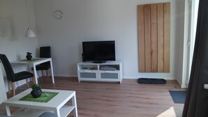 Een moderne woonkamer met een flatscreen televisie op een witte tv-meubel, een eethoek met een witte tafel en zwarte stoelen, en een houten garderobe tegen de muur.