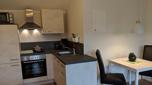 Een kleine keuken met een fornuis, oven en wastafel, een eettafel met een stoel, en een kleine potplant.