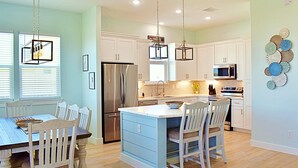 A kitchen with white cabinets, a stainless steel refrigerator, a microwave, and a large island with chairs. - Beach Please (Surfside Beach)