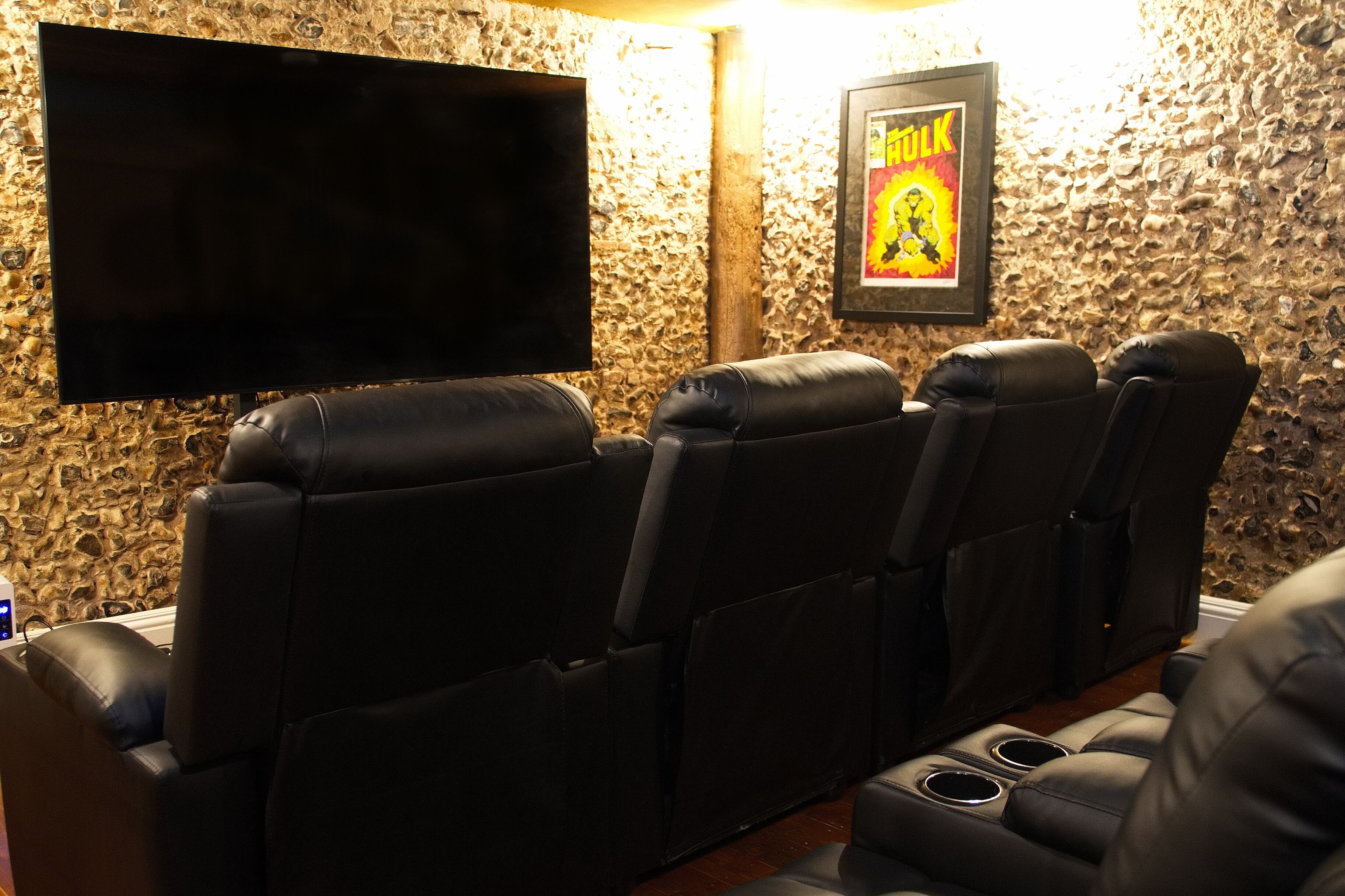 A home theater with four black leather recliner seats, a flat-screen TV mounted on the wall, and a framed poster on the wall.