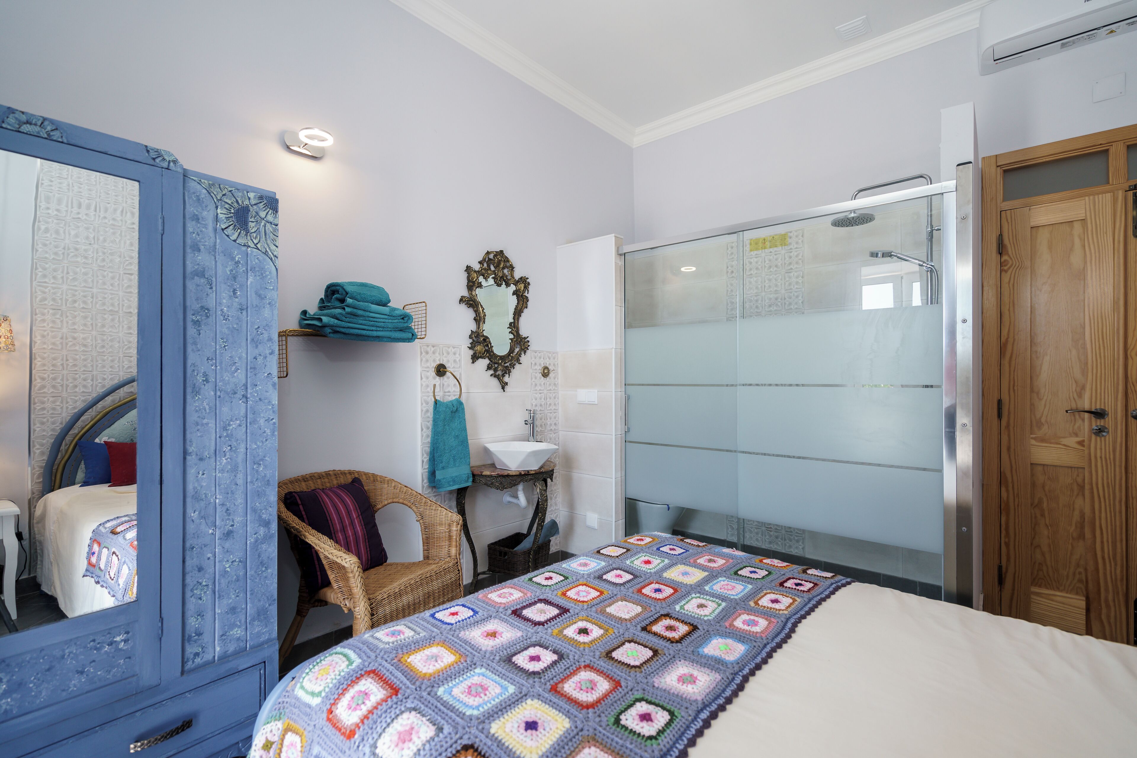 Une chambre à coucher comprenant un lit, un miroir, une chaise en osier, un lavabo et un espace douche.