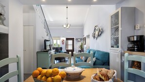 Een eetgedeelte met een houten tafel, een mand sinaasappels, een broodkom en een blauwe bank op de achtergrond.