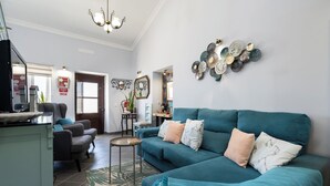 Een woonkamer met een blauwgroene bank, een bijzettafeltje en een kamerkleed. Er is een keuken met een koelkast en een magnetron.