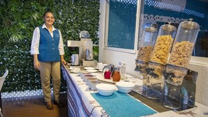 Montaje de bufé con gran variedad de alimentos y bebidas, una persona de pie a un lado y un fondo decorativo.
