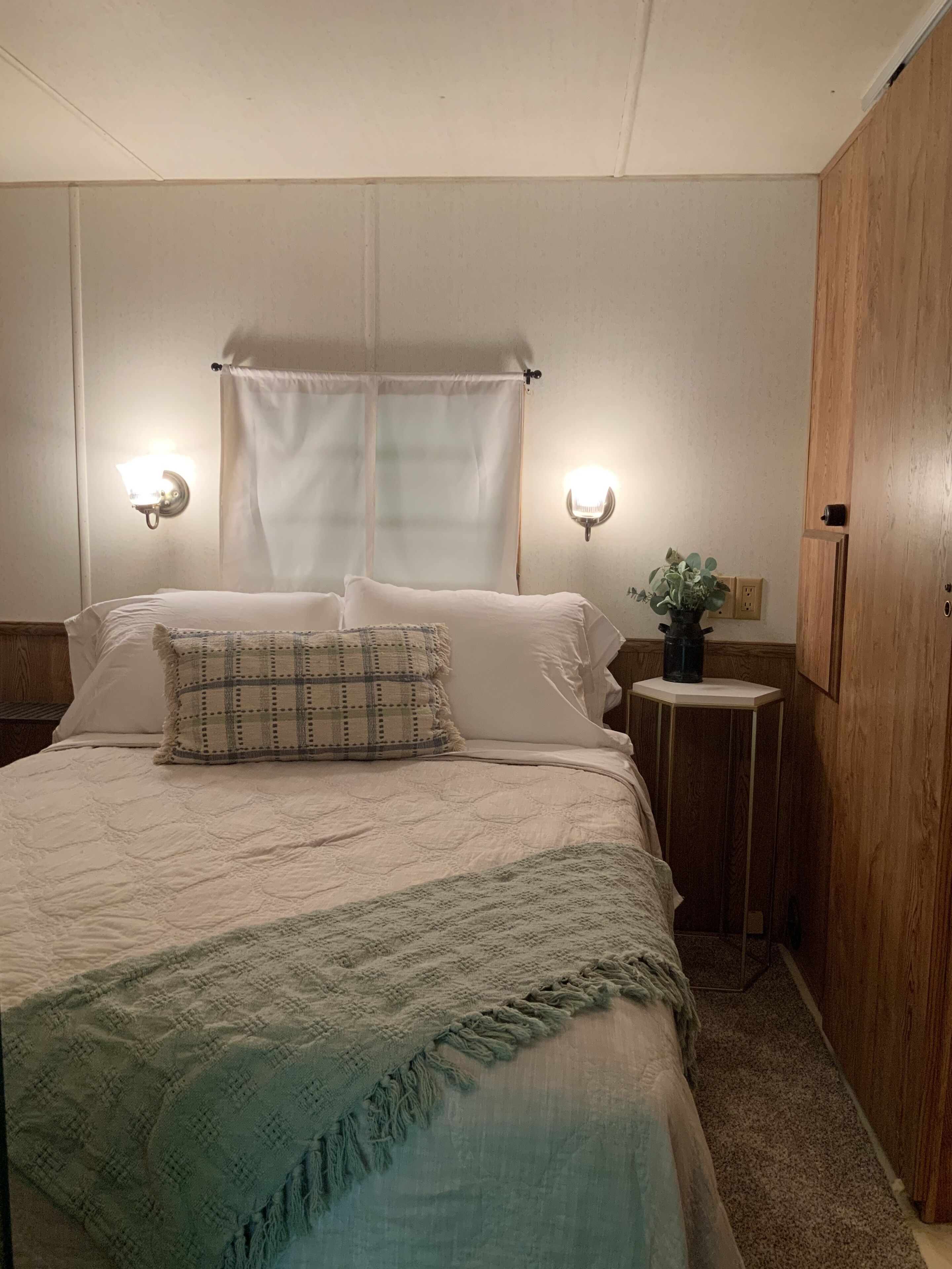 A neatly made bed with a plaid pillow, two wall-mounted lights, a small side table with a plant, and a wooden wardrobe.