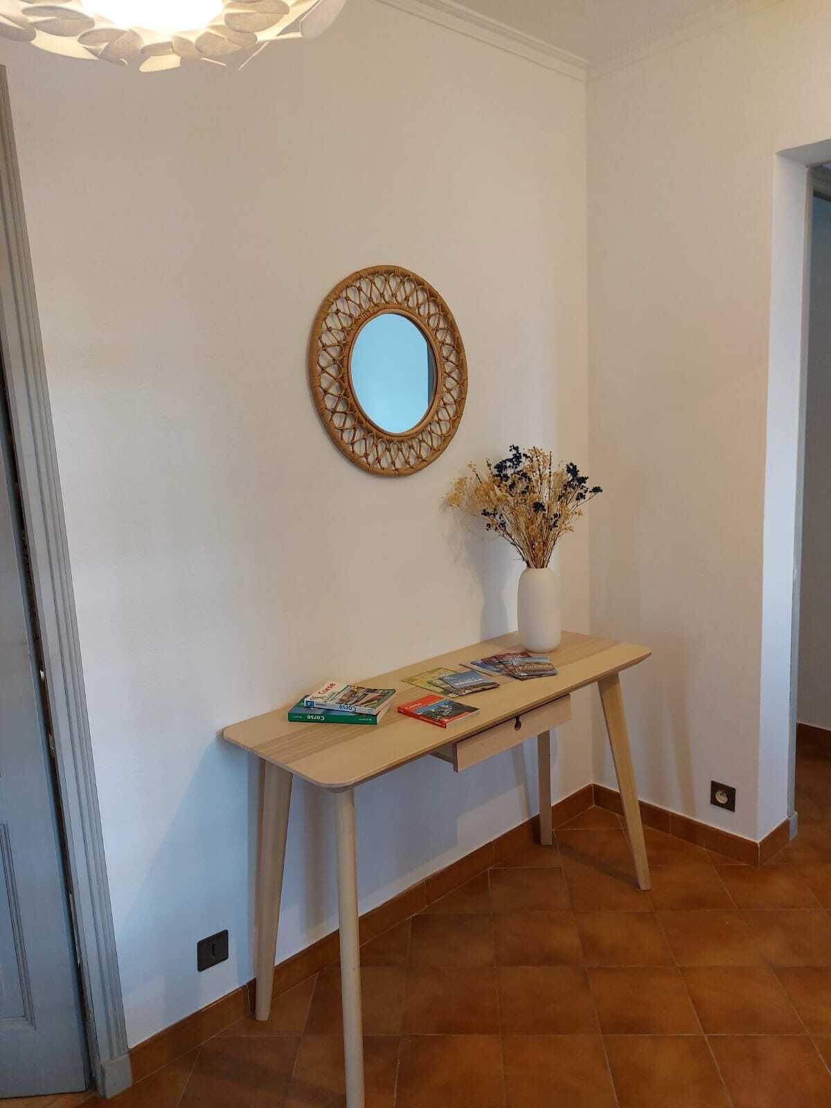 Un pasillo con una consola de madera que exhibe libros y un jarrón con flores secas. Un espejo redondo con marco tejido cuelga de la pared sobre la mesa.
