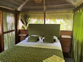Una cama con dosel estampado y almohadas, mesitas de noche de madera y vista a la vegetación a través de una ventana.