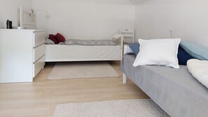 A small, well-lit room with a bed, a desk, and a chair. There is a white dresser and a small table with a lamp. The room has wooden flooring and a white ceiling. - Gemütliche Ferienwohnung in Ruhiger Lage (Sachsenheim)