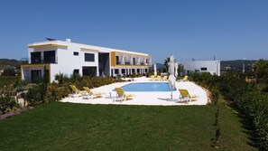 Um edifício moderno de dois andares com piscina, espreguiçadeiras e guarda-sóis ao redor.