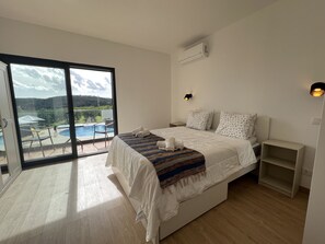 Quarto com cama, mesa de cabeceira, janela com vista para piscina e vegetação, e ar condicionado.