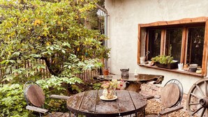 A rustic outdoor seating area with a round wooden table and metal chairs, surrounded by mulch and greenery. - Walnußhof im Spreewald (Schmogrow-Fehrow)