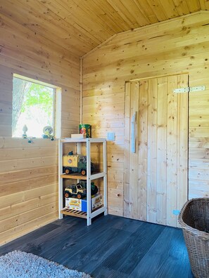 Ein Holzblockhaus-Interieur mit einem Regal voller Spielzeug-Lkw, einem Weidenkorb und einem Fenster mit Blick auf Bäume.