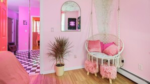 Una habitación con pared rosa, una silla colgante blanca con cojines rosados, una planta en maceta y suelo a cuadros.