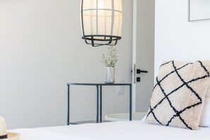 A bedroom with a bed, a bedside table with a vase of flowers, a hanging light fixture, and a decorative pillow.