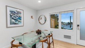 A dining area with a glass table and wooden chairs, a framed picture on the wall, and a window with a view of a parking lot. - The Sweetest Hill - 81 Walkscore (Seattle)