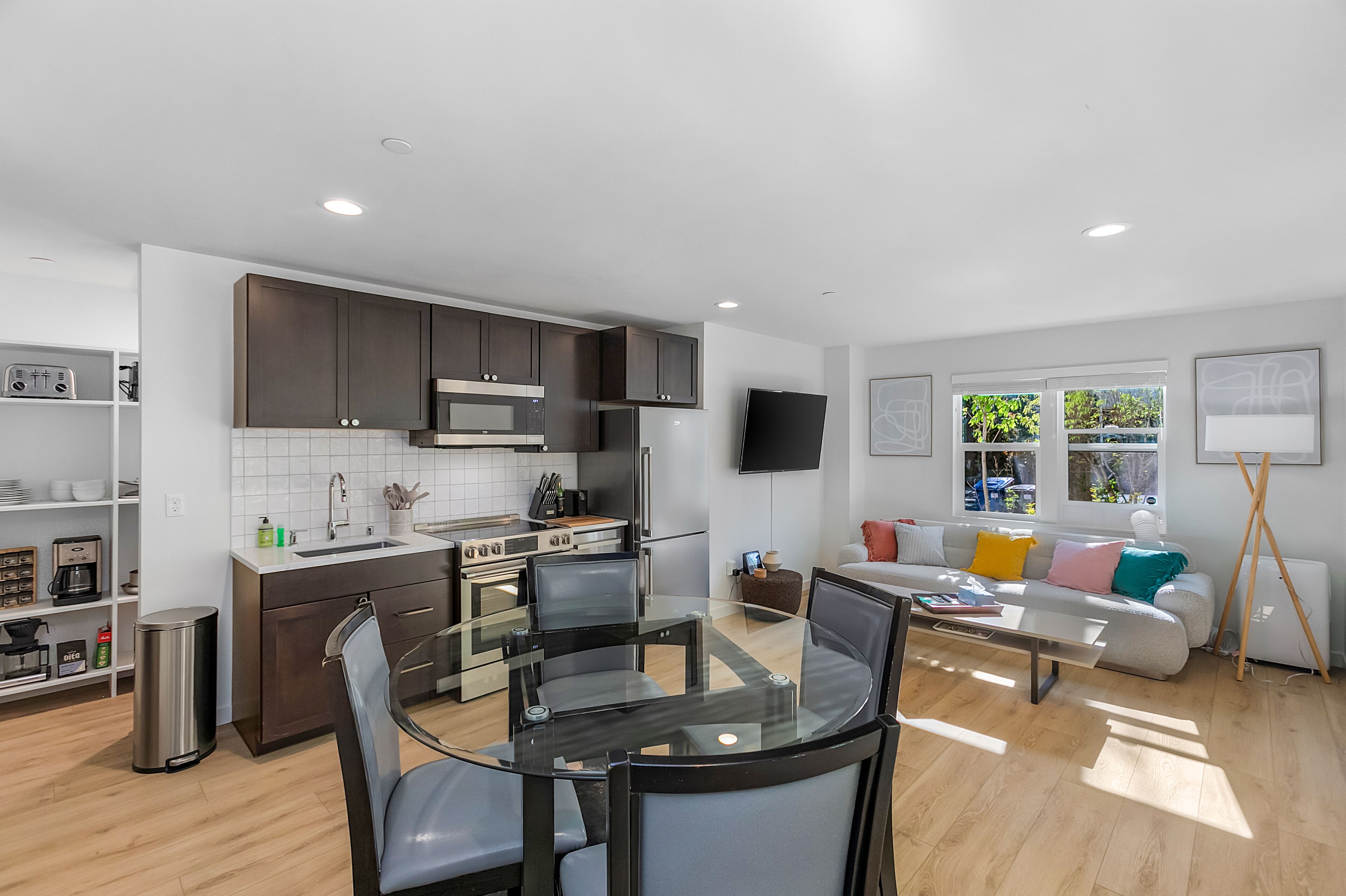 A modern kitchen with dark cabinets, a stainless steel range hood, and a built-in microwave. There is a dining area with a glass table and chairs, and a sofa with colorful cushions in the living room.