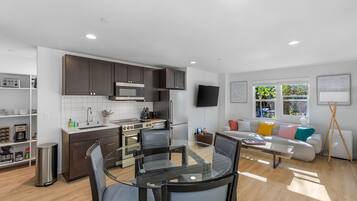 A modern kitchen with dark cabinets, a stainless steel range hood, and a built-in microwave. There is a dining area with a glass table and chairs, and a sofa with colorful cushions in the living room.