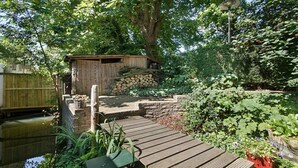 En træterrasse fører hen til et rustikt skur omgivet af frodig vegetation og en lille sø.