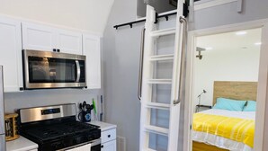 Uma cozinha compacta com micro-ondas, fogão e armários brancos. Uma porta de correr dá acesso a um quarto com cama e mesa de cabeceira.