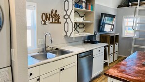 Cozinha moderna com armários brancos, eletrodomésticos de aço inoxidável e forno embutido. Há uma prateleira na parede com utensílios de cozinha e uma placa decorativa.