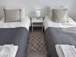 Two single beds with striped pillows and a throw blanket, a nightstand with a lamp and outlet, and a diamond-patterned rug complete this cozy retreat.