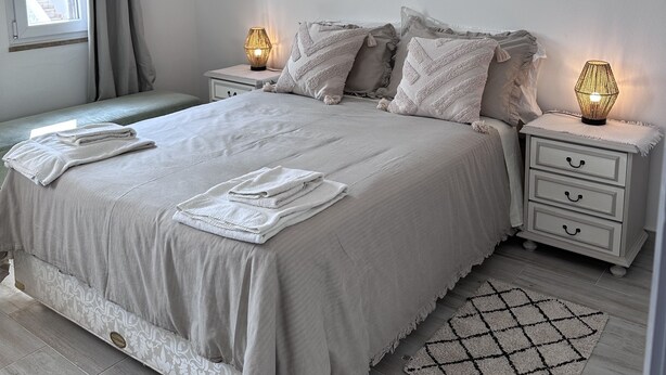 A modern bedroom with a large bed, bedside tables with lamps, a window with curtains, and a patterned rug. - Cosy refurbished 3 bedroom Vila in a quiet area 2km  from town center (Lagos)