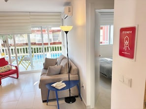 A living room with a sofa, a small table, a lamp, and a view of a pool outside.