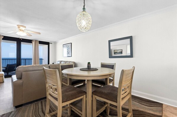 A room with a round table, four chairs, a ceiling fan, a hanging light fixture, and a view of the ocean through large windows. - Wind Drift South East 404 (Orange Beach)