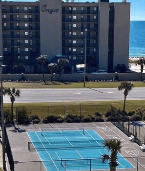 Cancha de tenis con piscina, palmeras y un edificio de hotel al fondo.