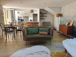 Un salon moderne avec un canapé, une table à manger et des chaises. Au fond, on aperçoit une cuisine équipée de placards et d’un plan de travail.