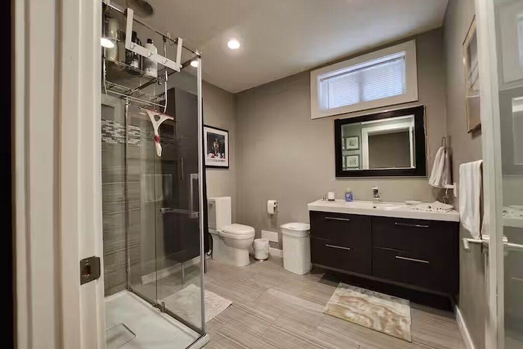 Un baño moderno con cabina de ducha de vidrio, un inodoro, un lavabo con espejo y un mueble de tocador de color oscuro.