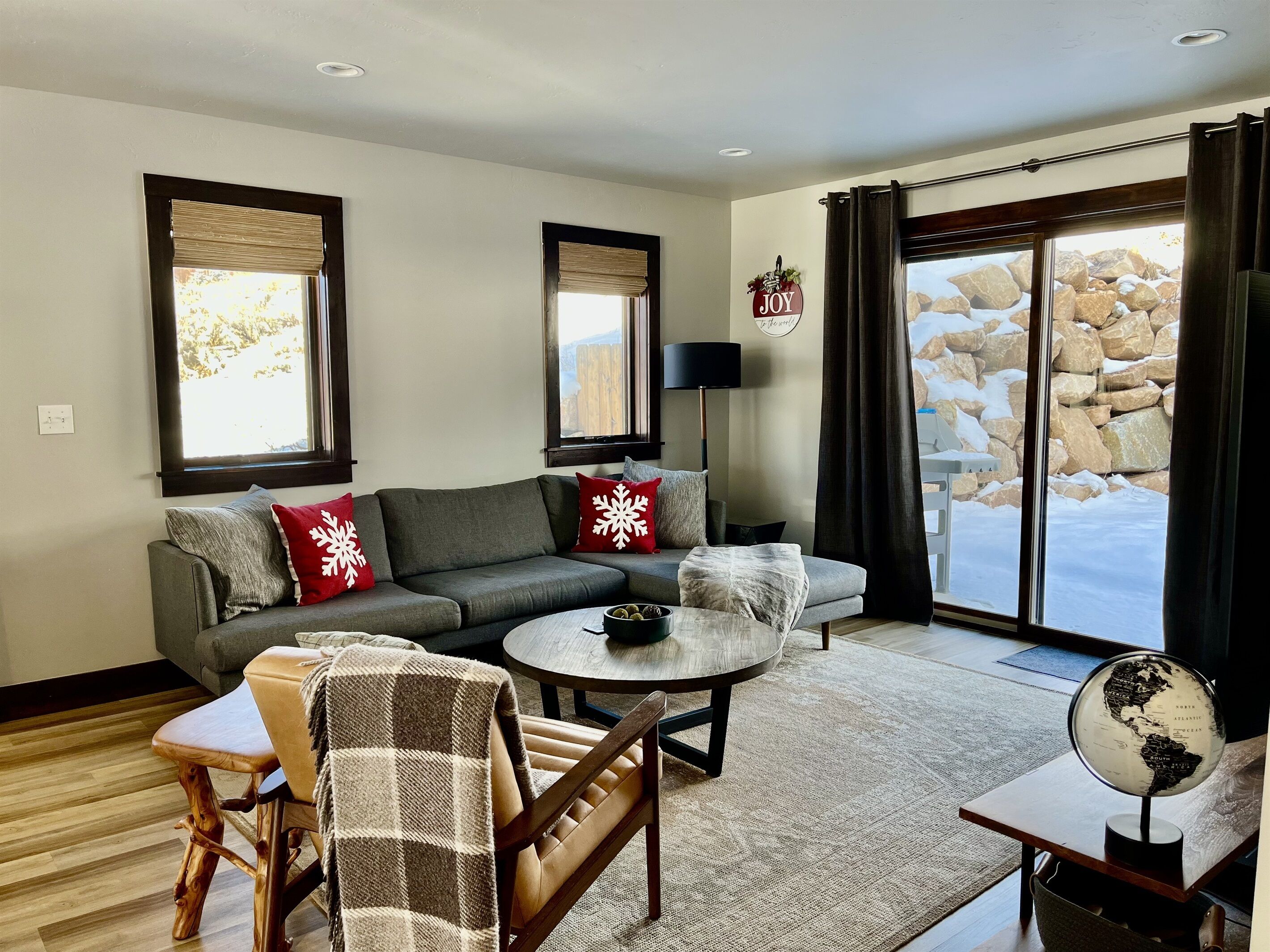 A cozy living room with a grey sofa, red and white snowflake pillows, a wooden chair, a small round table, and a globe on a side table.