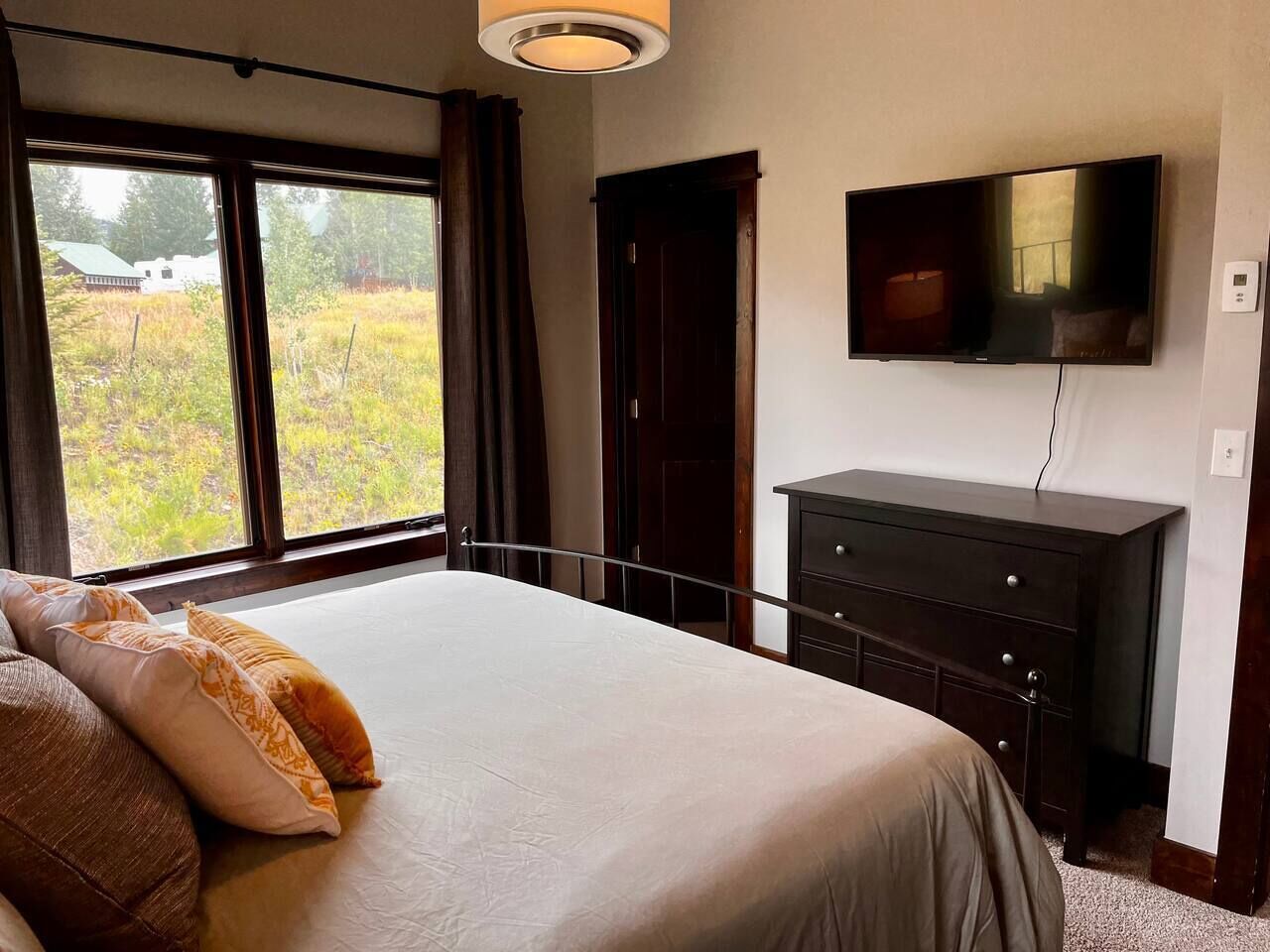 A bedroom with a large bed, a dresser, a television on the wall, and a window with a view of greenery.