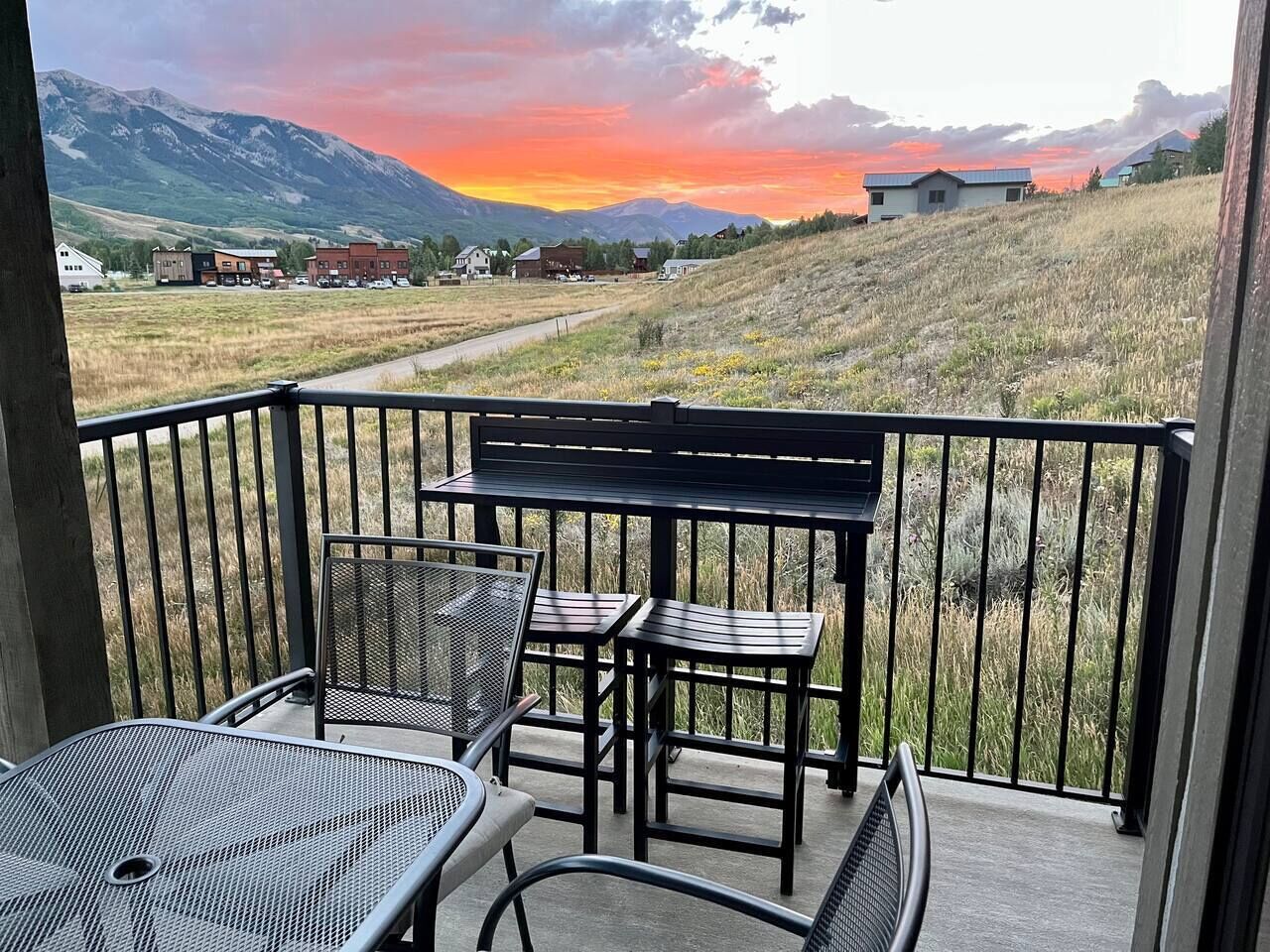 A balcony with a metal table and chairs, overlooking a grassy field and mountains during sunset.