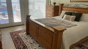 A bedroom with a wooden bed, a ceiling fan, a large window with blinds, a view of the backyard, and a painting on the wall. - Poolside Paradise on Cross Lake (Shreveport)