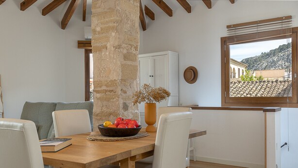 A dining area with a wooden table, white chairs, a bowl of fruit, and a view of a mountainous landscape through a window. - Townhouse with puig de Maria view by home villas 360 (Pollensa)
