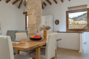 Uma área de refeições com uma mesa de madeira, cadeiras brancas, uma cesta de frutas e vista para uma paisagem montanhosa pela janela.