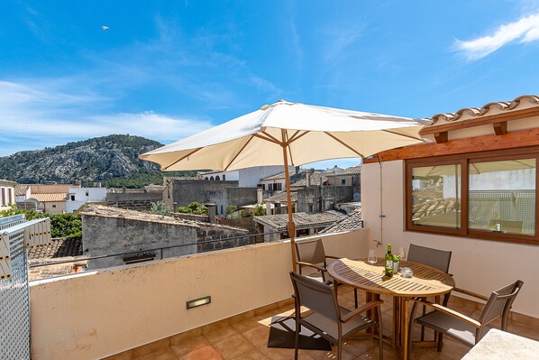 A rooftop terrace with a table and chairs, a large umbrella, and a view of the cityscape and mountains.