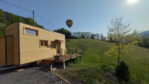 Eine kleine Holzhütte mit einer Terrasse, im Hintergrund ein Heißluftballon und ein grasbewachsener Hang.
