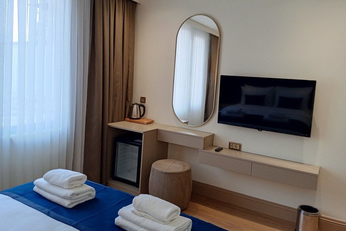 A hotel room with a bed, a TV, a mirror, a small table with a kettle, and a window with curtains.