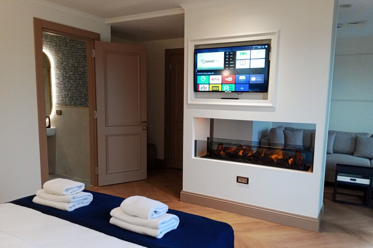 A hotel room with a bed, a TV mounted on the wall, a fireplace, and a sofa.